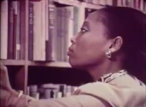 The still from 'Job Service Nebraska - Book Store' features a woman reaching for a book on a shelf, highlighting her focused expression. The setting is clearly a library or bookstore, with shelves filled with books in the background. The color tones have a vintage look typical of 16mm film, contributing to a warm, nostalgic atmosphere. This visual emphasizes the importance of education and resources available through the Job Service Nebraska initiative.