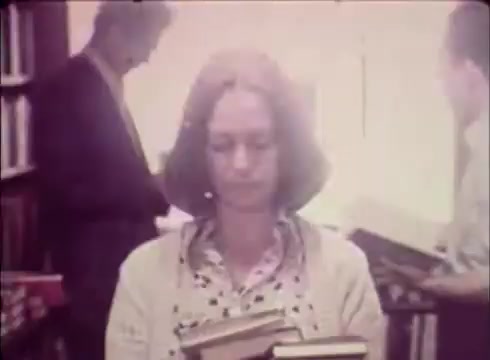 The 16mm still from 'Job Service Nebraska - Book Store' depicts a woman in a bright, softly lit bookstore, holding several books while looking contemplative. In the background, a man is seen browsing, contributing to the atmosphere of a quiet, literary space. The overall tone suggests a focus on education and resource access, typical of job service themes.