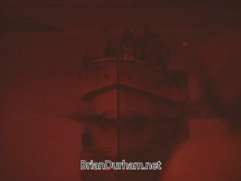 The still from the 'Columbian Fathers Derby NY PSA - 1970's' features a dimly lit scene dominated by a reddish hue, which creates a somber and reflective atmosphere. In the foreground, a boat appears to be moored or gently drifting on the water, with reflections visible on the surface. The overall composition conveys a sense of contemplation and purpose, aligning with the message of following a calling, as highlighted in the text about Jesus inviting his disciples to become 'fishers of men.' The visual elements support the theme of commitment to a spiritual journey.