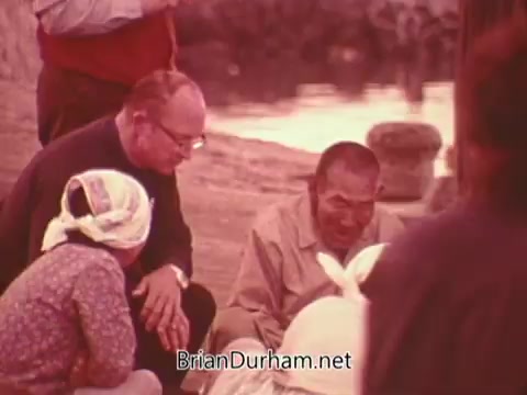 The still from the 'Columbian Fathers Derby NY PSA - 1970's' depicts a group of individuals engaged in conversation, likely in a setting that highlights community and connection. The scene features a priest or clergy member interacting with two other figures, one of whom appears to be a woman wearing a headscarf, while another man is seated and seemingly engaged in a dialogue. The setting suggests a warm, pastoral environment, possibly emphasizing outreach or missionary work. The text included references a biblical call to discipleship, inviting young individuals to consider a commitment to the Catholic priesthood. Overall, the image evokes themes of faith, community, and vocational dedication.