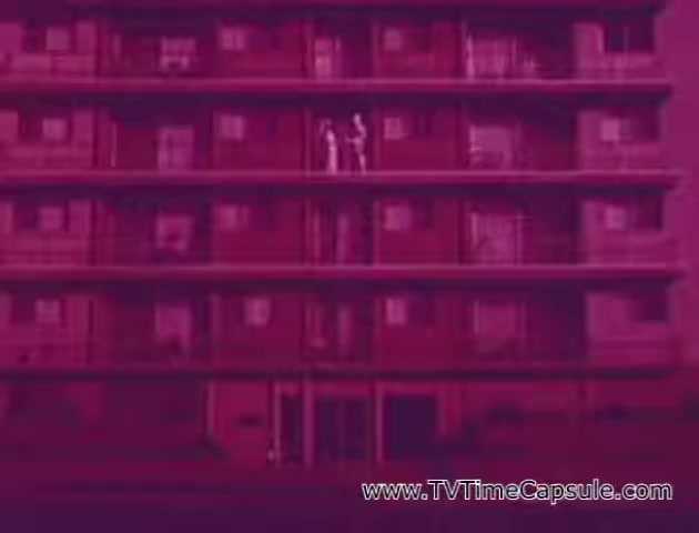 The still from the 'Lady Scott' commercials features a stylized, vibrant magenta backdrop of a multi-story building. Two figures, likely representing characters in the ad, are positioned on a balcony, enhancing the visual interest of the scene. The overall aesthetic suggests a playful and colorful marketing approach by the Scott Paper Company to promote their tissue and paper products. The setting and design seem aimed at capturing attention through bold colors and patterns.