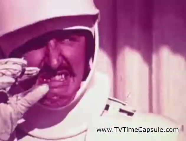 The still from the 'Lady Scott' commercials features a man in a futuristic, possibly space-themed outfit, complete with a helmet. He appears to be grimacing, possibly in an exaggerated or comedic manner. The colors are muted with a pinkish tint, creating a retro aesthetic typical of older commercials. This image likely aims to showcase the product in a lighthearted or humorous context. The overall vibe suggests a playful marketing approach by the Scott Paper Company to highlight their tissue and towel products.