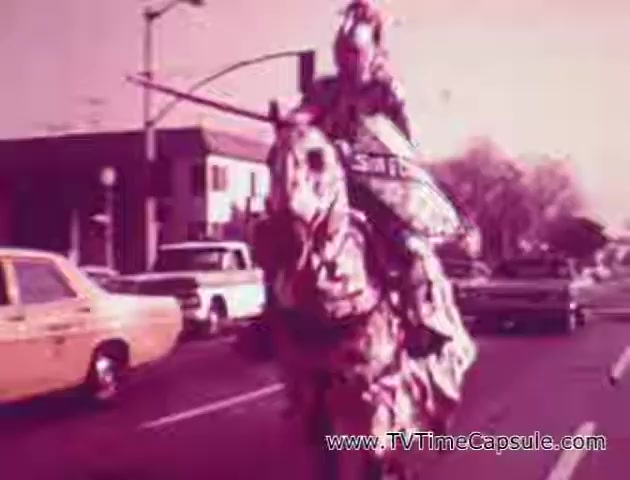The still from the 'Lady Scott' commercials features a whimsical scene set on a street, with a person dressed in a costume riding a horse. The rider appears to be adorned in colorful, patterned tissue-like material, showcasing the playful spirit of the advertisement. The background includes vintage cars, adding a classic touch. The overall color palette has a pinkish hue, enhancing the nostalgic vibe of the era. This promotional approach reflects the creative marketing strategies used by the Scott Paper Company to highlight their tissue products.