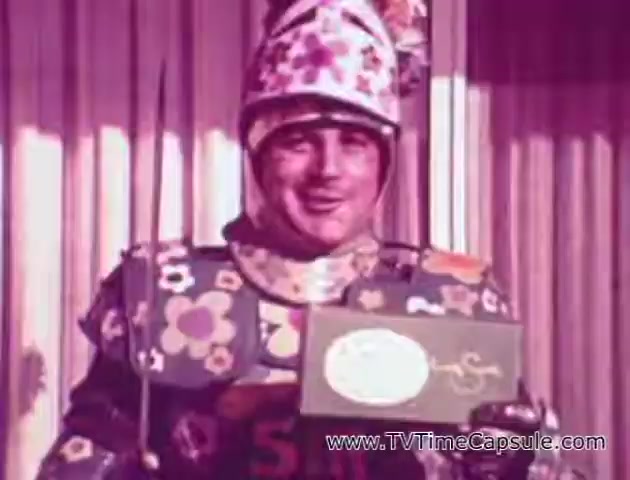 The still from the 'Lady Scott' commercials features a man in a whimsical costume adorned with colorful flower patterns. He is smiling and holding a box, presumably of the tissue or paper towel product. The background is simple, indicating a promotional setting. The vibrant colors and playful design emphasize the brand's effort to make household products more appealing.