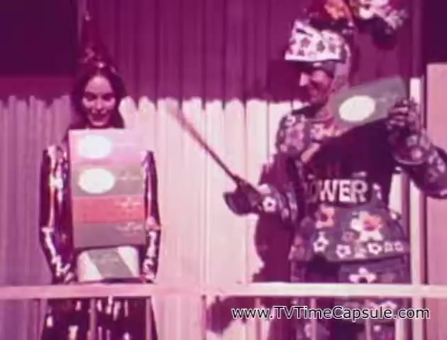 The still features a vibrant, theatrical setting with two characters: a woman in a colorful outfit and a man dressed in a costume adorned with patterns resembling tissue packaging. The man holds a pointer, emphasizing a product, likely a type of tissue or paper towel, while the woman displays another product box. The scene is lively and playful, showcasing the marketing style of the era for Scott Paper Company's 'Lady Scott' line.