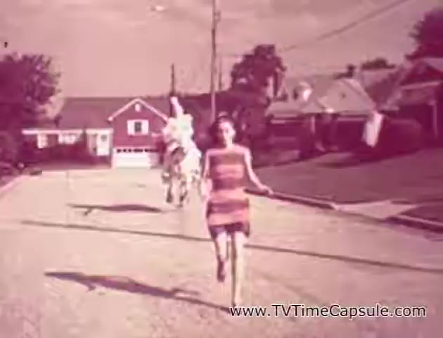 The still from the 'Lady Scott' commercials features a young girl running down a suburban street, seemingly in an early to mid-20th-century setting. She wears a striped dress and is followed by a person on a horse, who appears to be dressed in a whimsical, playful manner. The scene is tinted in warm pink hues, contributing to a nostalgic atmosphere. Houses line the street, with laundry hanging outside, reflecting a quaint neighborhood vibe. The overall composition is bright and fun, aiming to engage viewers with a sense of joy and playfulness associated with the Lady Scott brand.