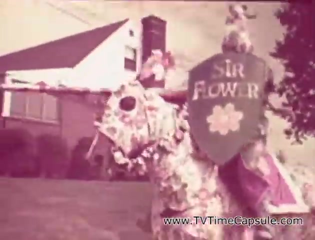 The still from the 'Lady Scott' commercials features a whimsical scene with a knight, adorned in floral decorations, riding a similarly decorated horse. The knight holds a shield with 'Sir Flower' written on it, suggesting a playful theme centered around flowers and nature. In the background, a suburban home is visible, enhancing the quaint, nostalgic vibe typical of mid-century advertisements. The colorful patterns and textures of the materials hint at the product's focus on bringing fun and creativity to everyday items like tissues and paper towels.