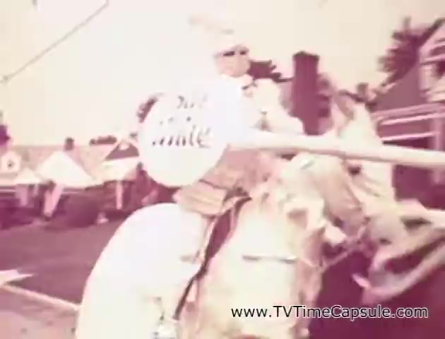The still from the 'Scott Towel 'Lady Scott'' commercials features a person riding a horse, holding a large sign with the text 'Lady Scott.' The scene appears to be set in a lively outdoor environment, potentially a fair or parade, with various colorful tents in the background. The commercial likely aimed to highlight the brand's products in an engaging, promotional manner, using a playful approach to attract attention. The overall aesthetic has a vintage vibe, characteristic of mid-20th century advertising.