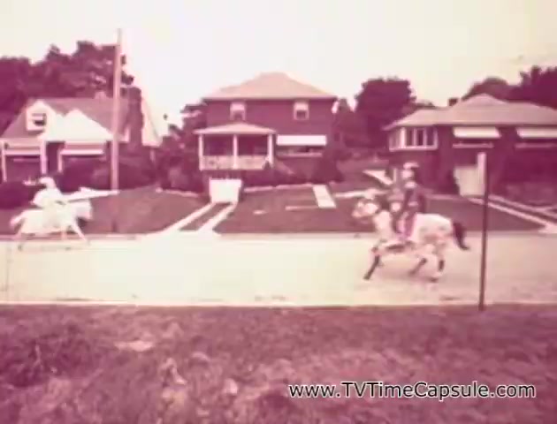The still from the 'Lady Scott' commercials features a whimsical scene set on a suburban street. Two individuals are riding horses, with one horse appearing white and the other colorful. The background includes quaint houses and green lawns, enhancing a nostalgic aesthetic. The overall tone seems lighthearted, likely aimed at showcasing the fun and versatility of the Lady Scott product line in a unique way. The commercial likely emphasizes creativity in home care with a playful visual narrative.