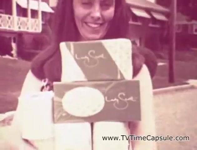 The still from the 'Scott Towel 'Lady Scott'' commercials features a woman smiling while holding various products, including packages of tissue and paper towels. The packaging is decorated with colorful patterns, emphasizing the brand's effort to make everyday items visually appealing. The setting appears to be a residential area, adding a relatable context to the advertisement. Overall, the image conveys a cheerful and lighthearted approach to promoting household products.