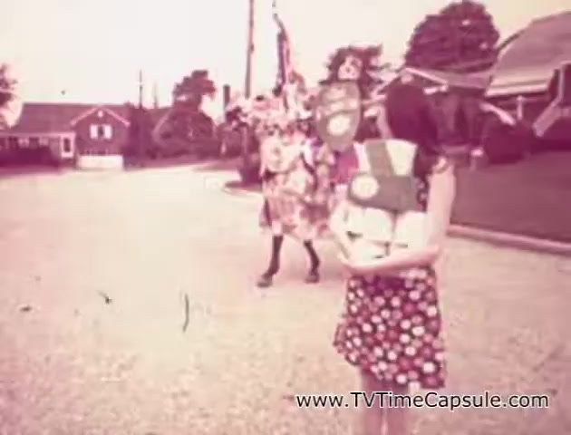 The still depicts a scene from a vintage 'Lady Scott' commercial featuring a young woman in a floral dress holding packages of tissue products, including paper towels and toilet paper. In the background, a performer dressed in a colorful costume, possibly a clown or mascot, adds a playful element to the scene. The setting appears to be a suburban street, with houses visible, creating a lighthearted and nostalgic atmosphere typical of mid-20th-century advertising. The overall color tone is faded, enhancing the retro aesthetic.