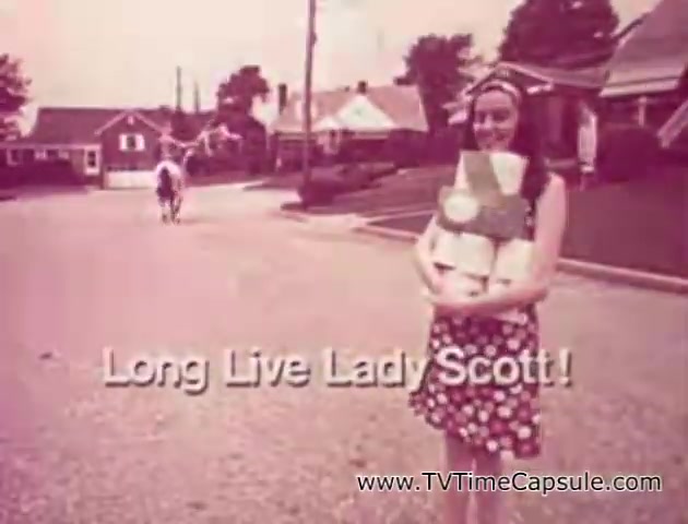 The still features a woman dressed in a floral-patterned dress and a headband, smiling as she carries stacks of 'Lady Scott' paper products, which include tissue and paper towels. She stands on a suburban street, with houses in the background, and a person on horseback can be seen in the distance. The text 'Long Live Lady Scott!' is prominently displayed at the bottom of the image, emphasizing the brand's celebration. The overall tone is nostalgic and cheerful, capturing a vintage advertising style.
