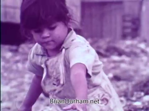 The still from the 16mm PSA features a young girl playing amidst debris, highlighting the challenges faced by children in need. The image conveys a sense of innocence and vulnerability against a backdrop of poverty, aligning with the message of the Smothers Brothers promoting the Foster Parents Plan. The context suggests an appeal for financial support to help overseas children.