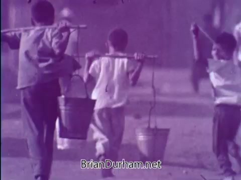 The still from the 1967 PSA by the Smothers Brothers shows three children walking together, each carrying a bucket suspended from a stick balanced on their shoulders. The children appear to be in a simple, outdoor setting, with a slightly faded or vintage color tone typical of film from that era. The scene reflects the theme of childhood and the aim of the PSA to raise awareness for the Foster Parents Plan.