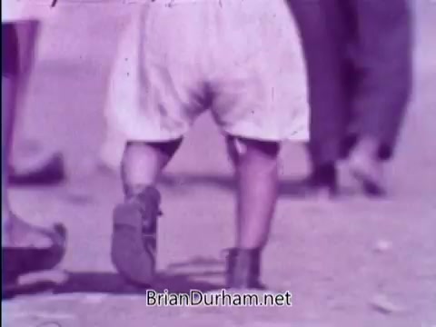The still from the 1967 PSA featuring the Smothers Brothers shows the lower half of a small child walking on a street. The child is dressed in light shorts and boots, with a focus on their footsteps. The background is somewhat blurred, indicating other people walking nearby, creating a sense of a bustling environment. The scene likely emphasizes the importance of supporting children in need through the Foster Parents Plan.
