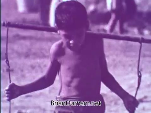 The still shows a young boy with a thin frame, appearing somber as he carries a large wooden stick across his shoulders. The image has a muted color tone, possibly giving a vintage feel typical of the 1960s. The scene emphasizes a poignant moment reflecting the struggles faced by children in need, aligning with the message of fostering support through the Foster Parents Plan.