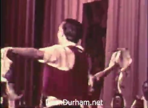 The still from the 'Pittsburgh Folk Festival PSA - 1968' showcases performers in traditional attire engaged in a dance. The scene appears to be set on a stage, with colorful drapes in the background. The event is presented by Robert Morris Junior College and highlights the 12th Pittsburgh Folk Festival, taking place at Civic Arena from June 7th to 9th. The performers seem to be celebrating cultural heritage through their dance movements.