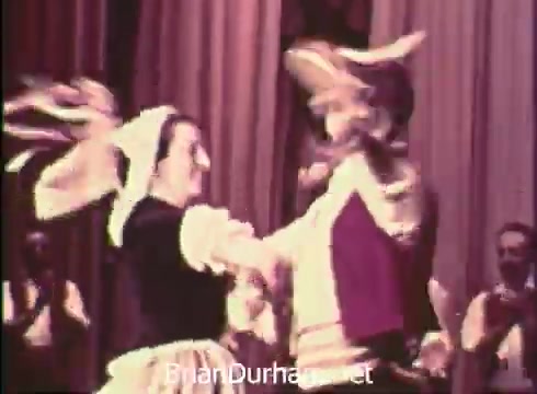 The still from 'Pittsburgh Folk Festival PSA - 1968' features a cultural dance performance. A male and female dancer are engaged in a lively folk dance, with traditional attire and dynamic movements. The background appears to have an audience and stage curtains, indicating a celebratory atmosphere for the festival, which took place at the Civic Arena on June 7th, 8th, and 9th.