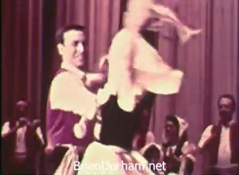 The still from 'Pittsburgh Folk Festival PSA - 1968' features a lively dance scene, showcasing traditional folk dancing. A male dancer, dressed in a vibrant outfit, appears to be spinning a female dancer who wears a decorative dress. The background includes other dancers, emphasizing a festive atmosphere. The context highlights the event hosted by Robert Morris Junior College at the Civic Arena from June 7th to 9th, celebrating cultural performances.