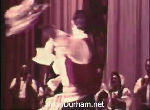 The still from 'Pittsburgh Folk Festival PSA - 1968' captures a lively performance scene. A dancer, dressed in traditional attire, spins energetically, showcasing vibrant movements. Behind them, a group of performers claps and supports the dance, enhancing the festive atmosphere. The event is promoted as part of the 12th Pittsburgh Folk Festival, taking place at Civic Arena from June 7th to 9th.