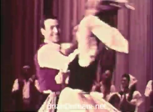 The still from the 'Pittsburgh Folk Festival PSA - 1968' depicts a lively folk dance performance. A male dancer, dressed in a vest, is spinning a female dancer wearing a traditional folk costume. Their engaging expressions and dynamic movements capture the spirit of the festival. The backdrop suggests a festive atmosphere, likely set in the Civic Arena during the event, which took place from June 7th to 9th.
