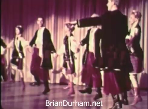 The still from 'Pittsburgh Folk Festival PSA - 1968' depicts a group of dancers performing on stage. They are dressed in traditional attire, showcasing a cultural dance. The backdrop features draped curtains, adding to the festive atmosphere. This moment highlights the celebration of folk culture as part of the festival organized by Robert Morris Junior College, which took place at the Civic Arena on June 7th, 8th, and 9th.