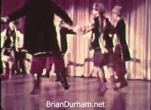 The still from the 'Pittsburgh Folk Festival PSA - 1968' features a group of dancers performing on stage. The dancers appear to be engaged in a traditional folk dance, showcasing colorful costumes and coordinated movements. The backdrop is likely a curtain, adding to the festive atmosphere of the event. The context refers to the 12th Pittsburgh Folk Festival, hosted by Robert Morris Junior College at the Civic Arena on June 7th, 8th, and 9th.