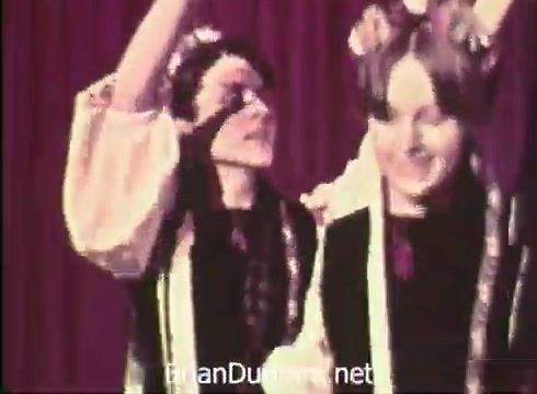 The still features two performers in traditional attire, likely representing ethnic folk culture, smiling and engaged in a lively moment. They are set against a red curtain backdrop, which enhances the festive atmosphere. This scene is a promotion for the 12th Pittsburgh Folk Festival, highlighting the event's focus on cultural celebration and community.