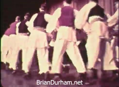 The 16mm still from the 'Pittsburgh Folk Festival PSA - 1968' shows a group of dancers performing in traditional attire. The dancers are arranged in a line, facing away from the camera, suggesting they are engaged in a lively folk dance. The setting appears to be on a stage, possibly during a performance at the Civic Arena, emphasizing the cultural celebration of the festival held from June 7th to 9th. The overall tone is vibrant and festive.