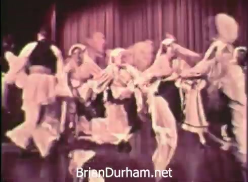 The still from the 1968 Pittsburgh Folk Festival PSA depicts a lively performance featuring dancers in traditional costumes. They are engaged in a group dance, showcasing cultural heritage and community spirit. The background features a stage curtain, suggesting a formal setting for the festival. The event is presented by Robert Morris Junior College and celebrates folk traditions.