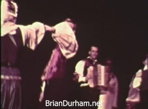 The still from the 'Pittsburgh Folk Festival PSA - 1968' depicts a lively performance featuring dancers in traditional attire. One dancer is gesturing with a piece of clothing, while another plays the accordion, creating a festive atmosphere. The context mentions the 12th Pittsburgh Folk Festival, hosted at the Civic Arena from June 7th to 9th, emphasizing the event's cultural significance and celebration of folk traditions.