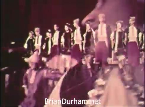 The still from the 'Pittsburgh Folk Festival PSA - 1968' features a group of performers dressed in traditional attire, likely representing various cultural backgrounds. They stand in formation, possibly preparing for a dance or musical performance, with an emphasis on vibrant costumes. The event is presented by Robert Morris Junior College and highlights the 12th Pittsburgh Folk Festival, taking place at the Civic Arena from June 7th to 9th, celebrating diverse folk traditions.
