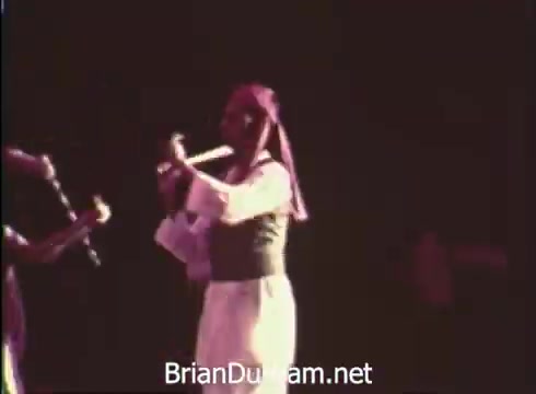 The still depicts a performer playing a flute, likely part of a dance or music ensemble at the 12th Pittsburgh Folk Festival, held at the Civic Arena in June 1968. The performer is dressed in traditional attire, showcasing the festival's celebration of cultural arts and folk traditions. The lighting emphasizes the stage environment, hinting at a vibrant festival atmosphere.