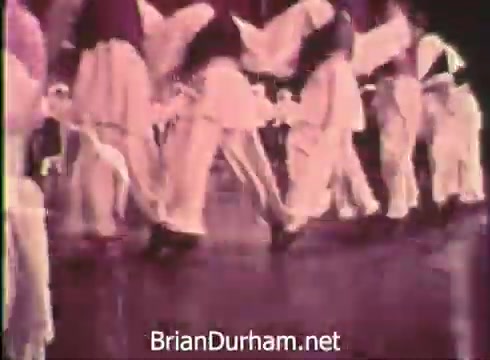 The still from 'Pittsburgh Folk Festival PSA - 1968' captures dancers in traditional attire performing together. Their costumes are characterized by flowing garments, and the scene emphasizes the movement of their feet on a polished floor. The setting conveys a lively atmosphere, likely showcasing cultural heritage during the festival held at the Civic Arena on June 7th, 8th, and 9th.