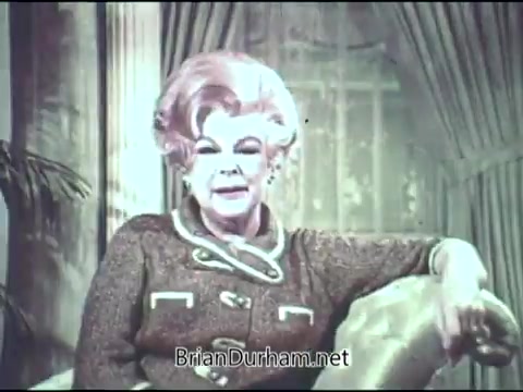 The still from the 1969 American Cancer Society PSA features Virginia Graham, a television personality, seated in a comfortable setting. She is dressed in a stylish, patterned outfit and speaks directly to the audience, conveying an important message related to cancer awareness. The background includes soft drapes and foliage, creating a warm, inviting atmosphere.