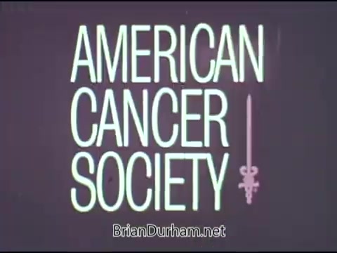 The still features the title 'AMERICAN CANCER SOCIETY' prominently displayed in a bold, stylized font. The text is accompanied by a decorative element resembling a sword, contributing to a serious and impactful visual. The overall color scheme is likely to be simple, enhancing legibility and focus on the message. This PSA likely aims to raise awareness about cancer during a critical time in public health discussions.