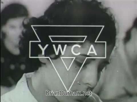 The still features a close-up of a young woman, highlighting her expression, with the YWCA logo prominently displayed in the foreground. The setting appears to involve a group, suggesting a focus on community or educational themes relevant to the YWCA's mission. The black-and-white presentation reflects a historical context, likely aimed at promoting awareness or services offered by the organization.