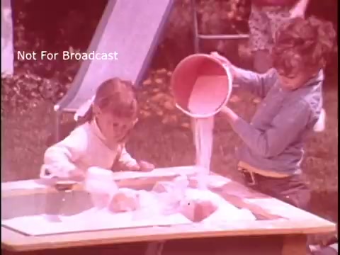 The still features two young children engaged in a playful activity outdoors. One child, dressed in a light sweater, is pouring a substance from a bucket onto a table, while the other, wearing a blue shirt, is focused on the table as well. The scene captures a sunny day, evoking a sense of childhood innocence and creativity, which is likely tied to the theme of product use in the Baggies food storage bag commercial.