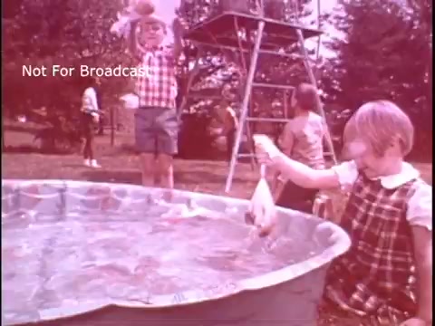 The still from the 'Baggies food storage bag commercial' features children playing outdoors near a small pool. One child, dressed in a checkered shirt and shorts, is joyfully holding up something, possibly related to the product. Another child sits by the poolside, examining or playing with an item in their hands. The scene conveys a playful and carefree summer atmosphere, emphasizing the fun and versatility of Baggies food storage bags in family activities. The color tone appears faded, typical of vintage commercials.