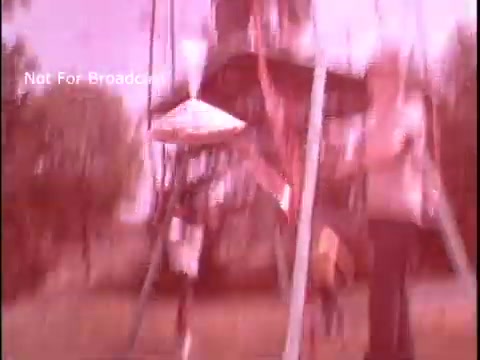 The still from the 'Baggies food storage bag commercial' features a nostalgic, slightly faded 16mm film aesthetic. In the image, a child swings on a playground swing set while an adult stands nearby, possibly engaging with them. The background suggests a sunny day in a suburban environment, emphasizing a family-friendly atmosphere. The focus appears to be on the playful interaction, aligning with the commercial’s theme of practicality and fun in food storage.