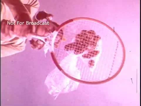 The still features a playful perspective from below a tennis racket, looking up. A hand is about to hit a Baggies food storage bag, which contains food items. The background has a whimsical, slightly pink hue, emphasizing the lighthearted nature of the commercial. The focus is on the action of the bag being 'hit,' suggesting the durability and fun aspect of using Baggies for food storage.