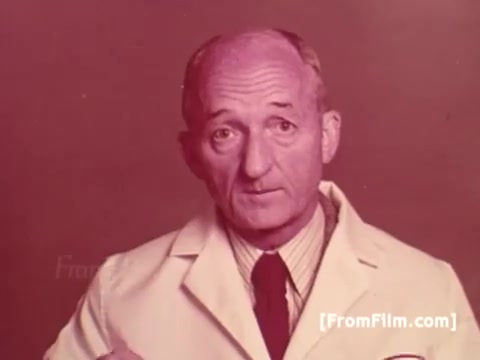 The still features Frank Perdue, dressed in a white lab coat and a tie, standing against a muted background. He appears serious and engaging, likely delivering a message about quality chicken production. This image captures the essence of 1970s advertising, emphasizing his tagline about the toughness required in chicken farming.