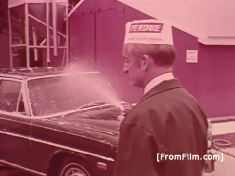 The still features Frank Perdue wearing a distinctive Perdue Chicken hat, standing next to a car being sprayed with water. The background is a solid color, possibly depicting a chicken processing or marketing setting. The context reflects the brand's focus on quality, with the tagline emphasizing the toughness required to produce a superior product. The visual style is characteristic of 1970s advertising, utilizing a straightforward approach to convey the message.