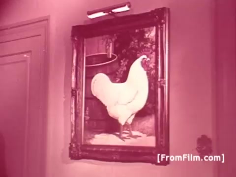 The still features a framed painting of a large white chicken, prominently displayed on a wall. The setting appears to be an indoor space, likely a home or barn, indicated by the decor and lighting. The image has a distinctive pinkish hue, typical of some vintage 16mm film prints. This scene reflects the light-hearted and humorous tone often found in Perdue Chicken commercials from the 1970s, emphasizing the brand's connection to quality and care in chicken farming.