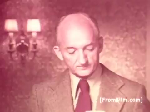 The still features Frank Perdue, a middle-aged man with a bald head, dressed in a suit and tie. He appears to be contemplating or reading something, with a thoughtful expression. The background is soft and muted, suggesting a domestic setting, possibly highlighting the warmth associated with his chicken brand. The overall tone reflects the vintage style of 1970s commercials, consistent with his tagline about the toughness needed to produce quality chicken.