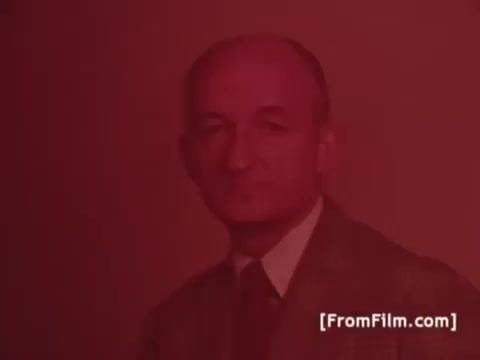 The still is from a 16mm print of a 1970s Perdue Chicken commercial featuring Frank Perdue. He is likely appearing in a promotional context, embodying the tagline, 'It takes a tough man to grow a tender chicken,' which emphasizes his hands-on approach to chicken farming. The overall aesthetic reflects the vintage style of the era's advertisements.