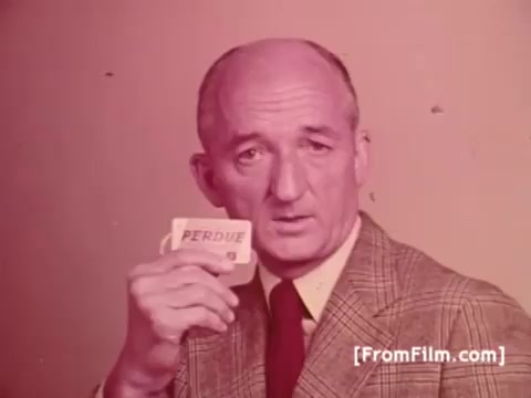 The still features Frank Perdue, the founder of Perdue Chicken, holding a card with the brand's name. He is dressed in a suit, conveying a serious demeanor that aligns with his tagline, 'It takes a tough man to grow a tender chicken.' The vintage aesthetic of the 1970s is evident, giving the image a nostalgic feel. The overall tone suggests both authority and trust in the quality of the product.