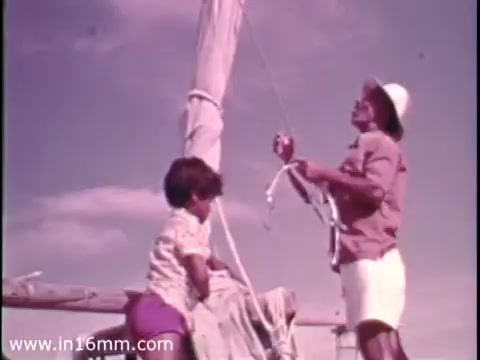 The still from the 1975 Christian Children's Fund PSA features a sunny outdoor setting with a man and a child engaged in activities involving a boat. The man, wearing a hat and shorts, appears to be handling ropes while the child, dressed in a light shirt and shorts, is sitting and watching. The colors are warm, reflecting the vintage style of the era. The scene emphasizes collaboration and mentorship, typical themes for such public service announcements.