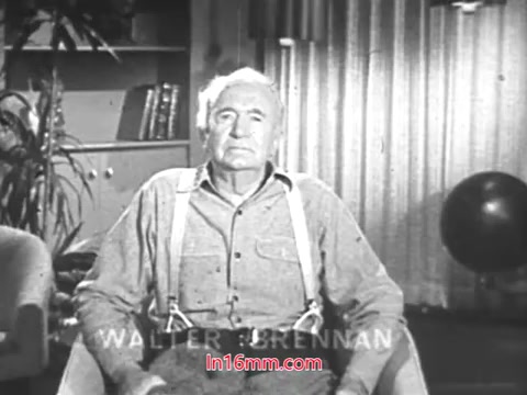 The still features Walter Brennan, an older man with a serious expression, seated in a living room setting. He wears a button-up shirt with suspenders, conveying a classic, nostalgic look. The background includes a plant and bookshelves, creating a warm, domestic atmosphere. This PSA promotes the tradition of using Christmas Seals, emphasizing their significance during the holiday season.