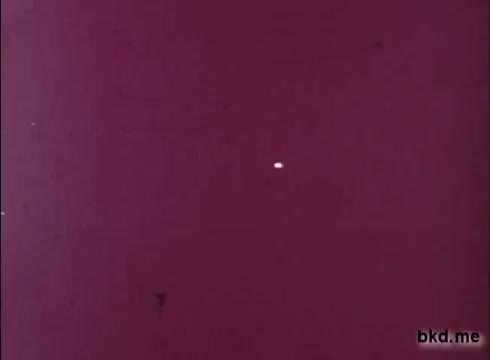 The still features a plain, reddish background with a small, white object seemingly floating in the center. The image likely reflects the vintage characteristics of a 16mm print, possibly showing signs of aging such as grain or scratches. The simplicity and color scheme align with aesthetic trends from the 1970s.